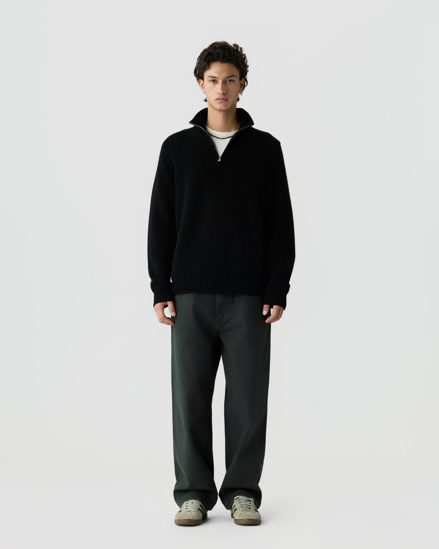 A young man stands against a plain light gray background wearing a black quarter-zip sweater, dark wide-leg pants, and light-colored sneakers.