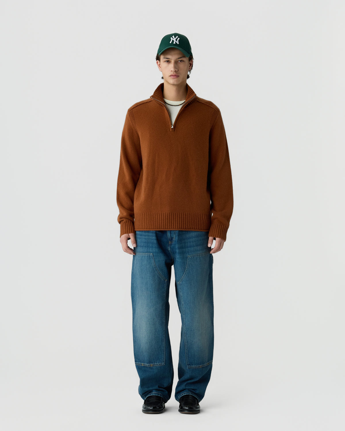 A young man wearing a brown quarter-zip sweater, blue jeans, black shoes, and a green baseball cap with a white logo stands against a plain white background.