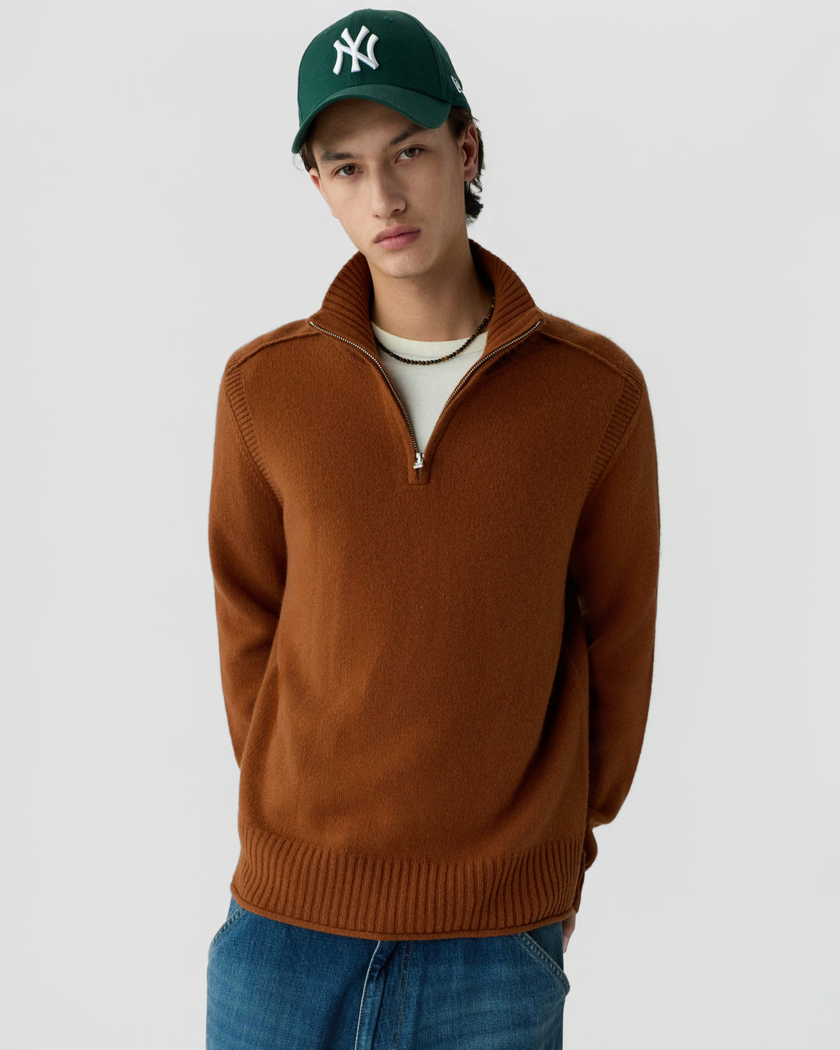 A young man wearing a brown quarter-zip sweater, blue jeans, and a dark green baseball cap with a white logo stands against a plain light background.