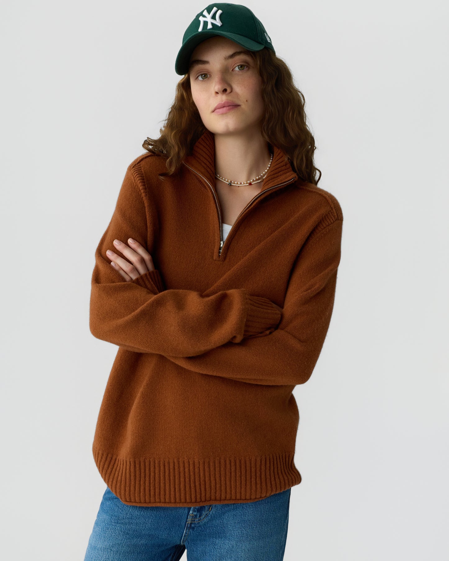 Young woman wearing a brown zip-up sweater and green baseball cap with crossed arms against a plain light background.