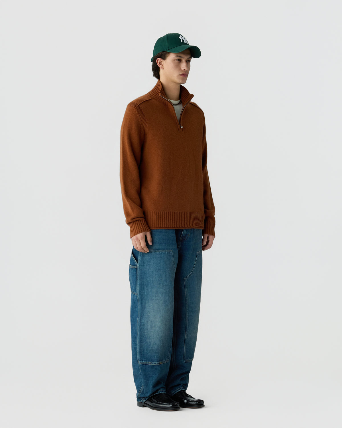 A young man wearing a brown half-zip sweater, loose-fitting blue jeans, black shoes, and a dark green baseball cap stands against a plain white background.
