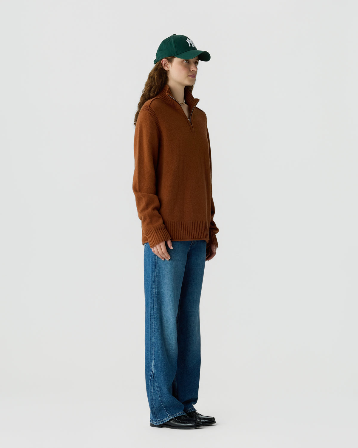 Woman wearing a brown zip-up sweater, blue wide-leg jeans, black shoes, and a dark green baseball cap standing against a plain white background.
