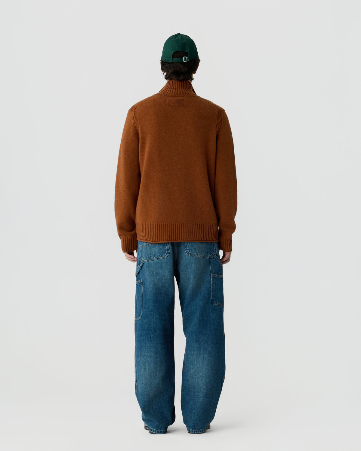 Person wearing a brown long-sleeve sweater and loose-fitting blue jeans, standing facing away against a plain light background, with a dark green cap on their head.