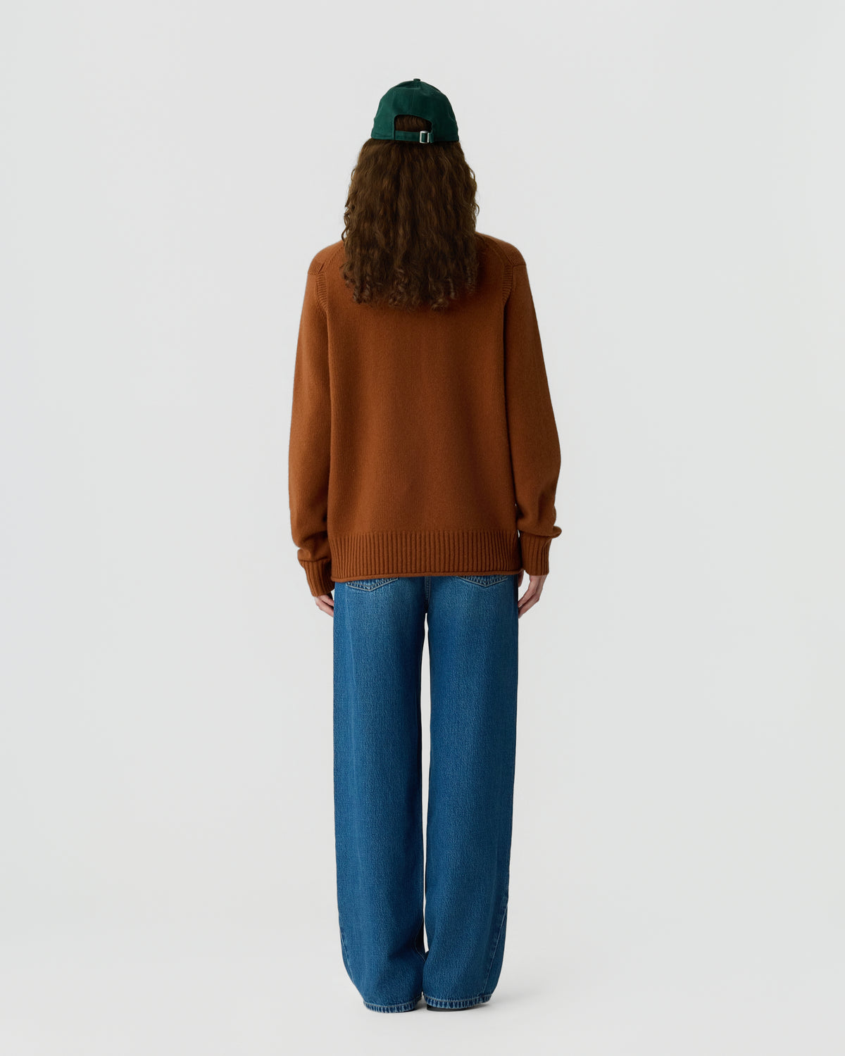 Person wearing a brown long-sleeve sweater, blue wide-leg jeans, and a dark green baseball cap, standing facing away against a plain light gray background.