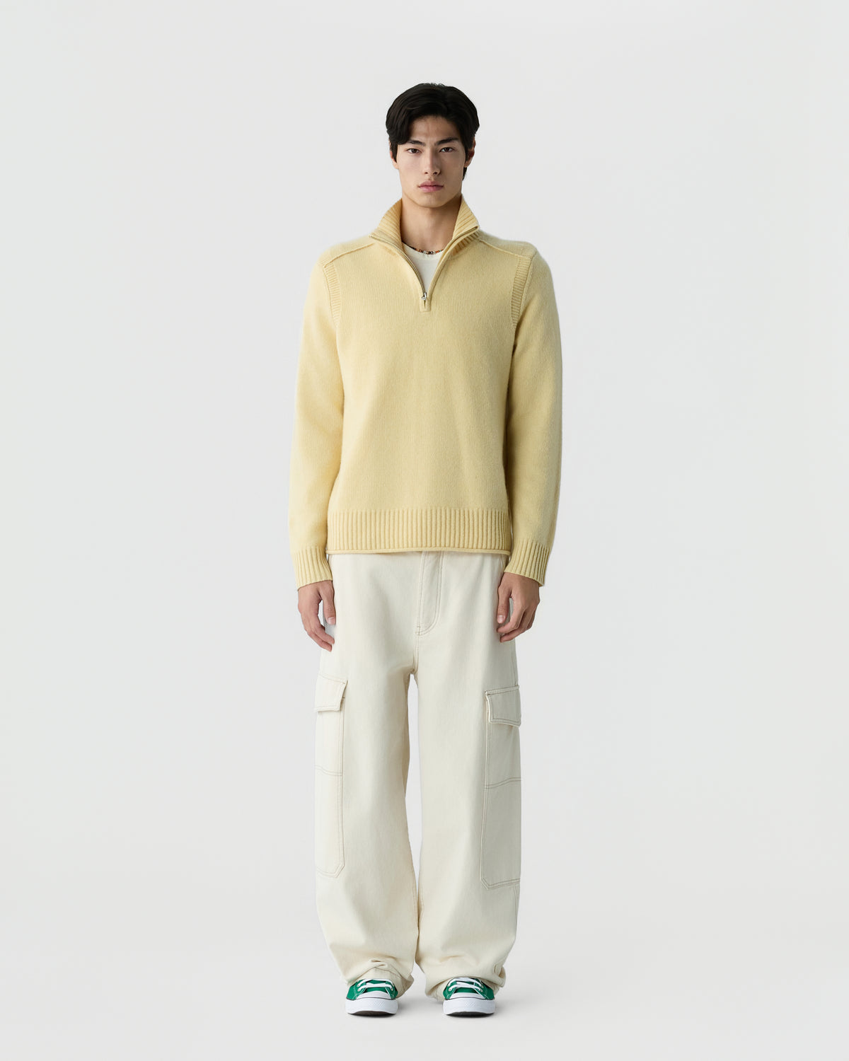A man stands facing forward wearing a light yellow zip-up sweater, off-white cargo pants, and green and white sneakers against a plain white background.