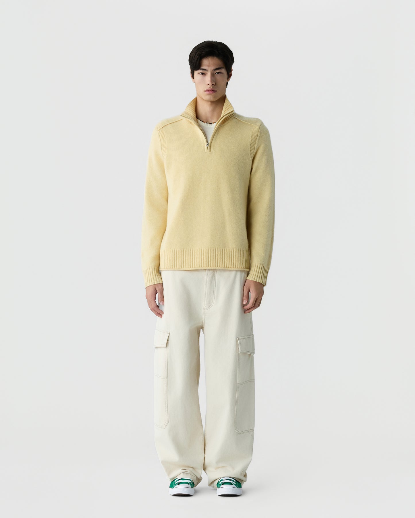 A man stands facing forward wearing a light yellow zip-up sweater, off-white cargo pants, and green and white sneakers against a plain white background.