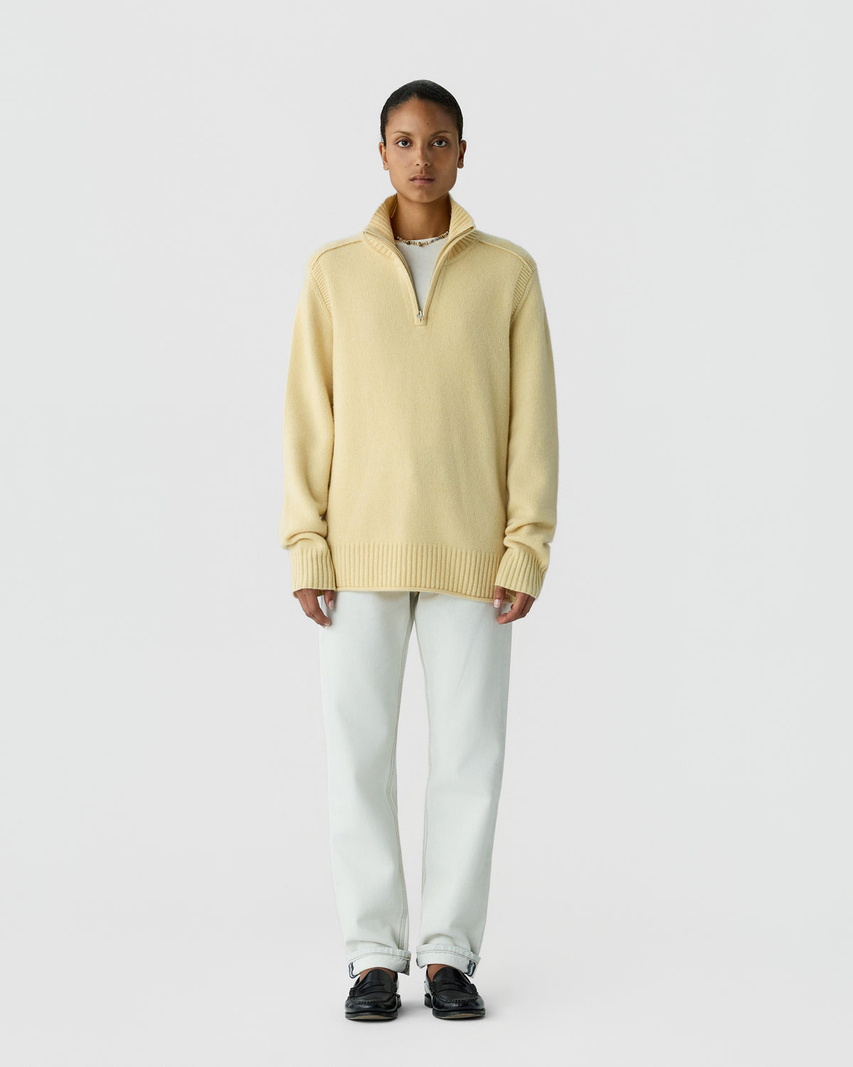 Person wearing a pale yellow oversized knit sweater with a quarter zipper and high collar, white pants, and black slip-on shoes standing against a plain white background.