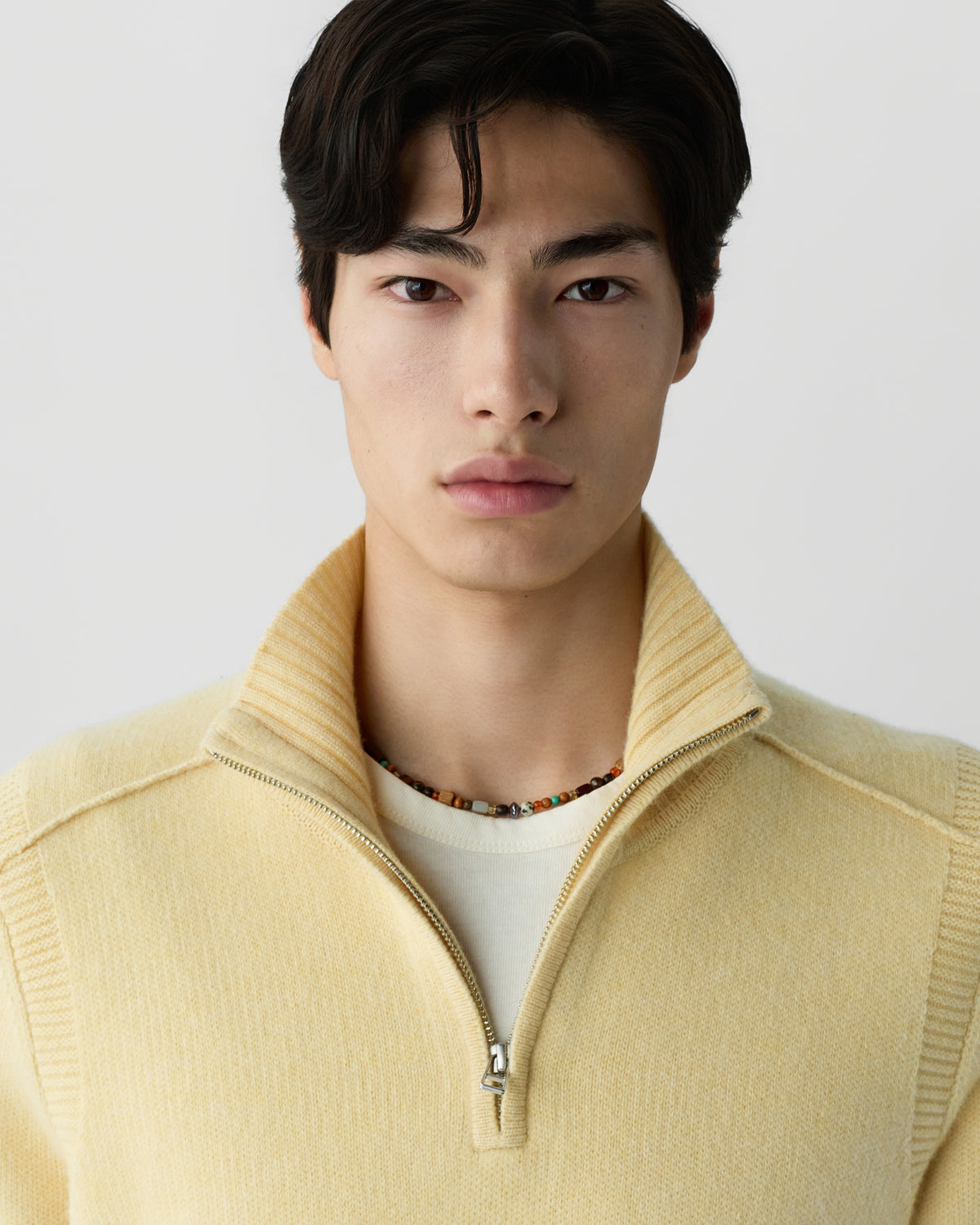 Young man with dark hair wearing a pale yellow half-zip cashmere sweater layered over a white shirt and a beaded necklace, facing forward against a neutral background.