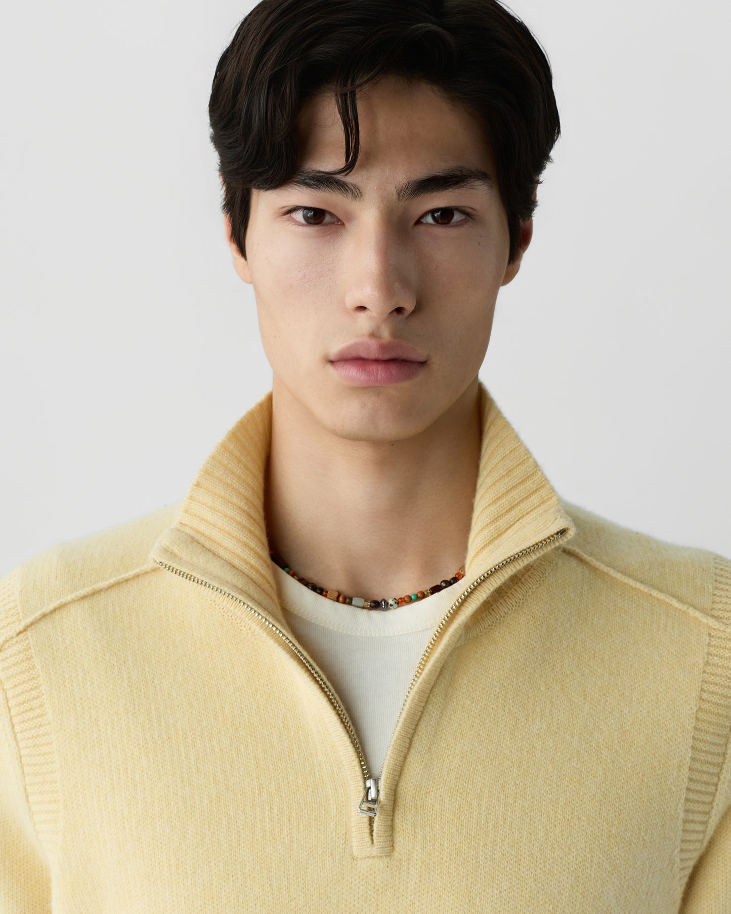 Young man with dark hair wearing a pale yellow half-zip cashmere sweater layered over a white shirt and a beaded necklace, facing forward against a neutral background.