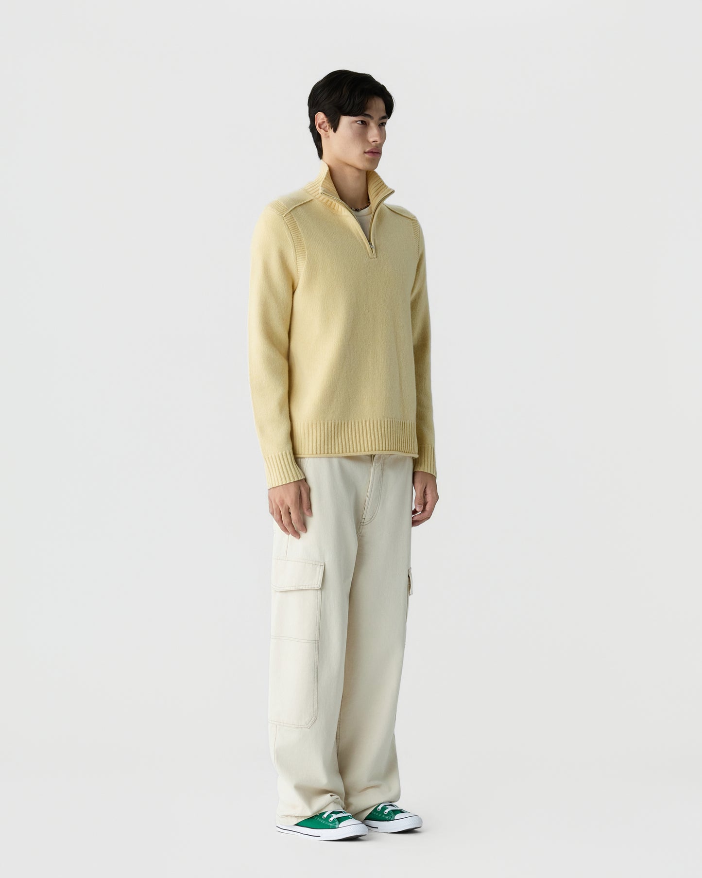 A man stands against a plain white background wearing a light yellow zip-up sweater with a high collar, cream cargo pants, and green sneakers with white soles.