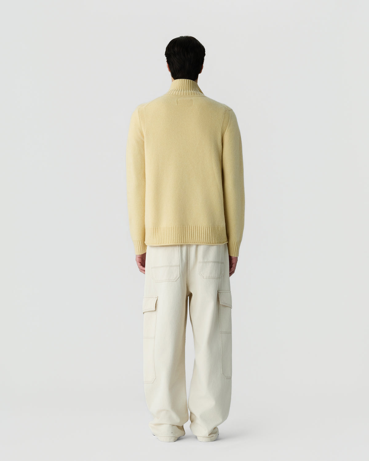 Rear view of a person wearing a light yellow cashmere turtleneck sweater paired with loose-fitting off-white cargo pants against a plain white background.