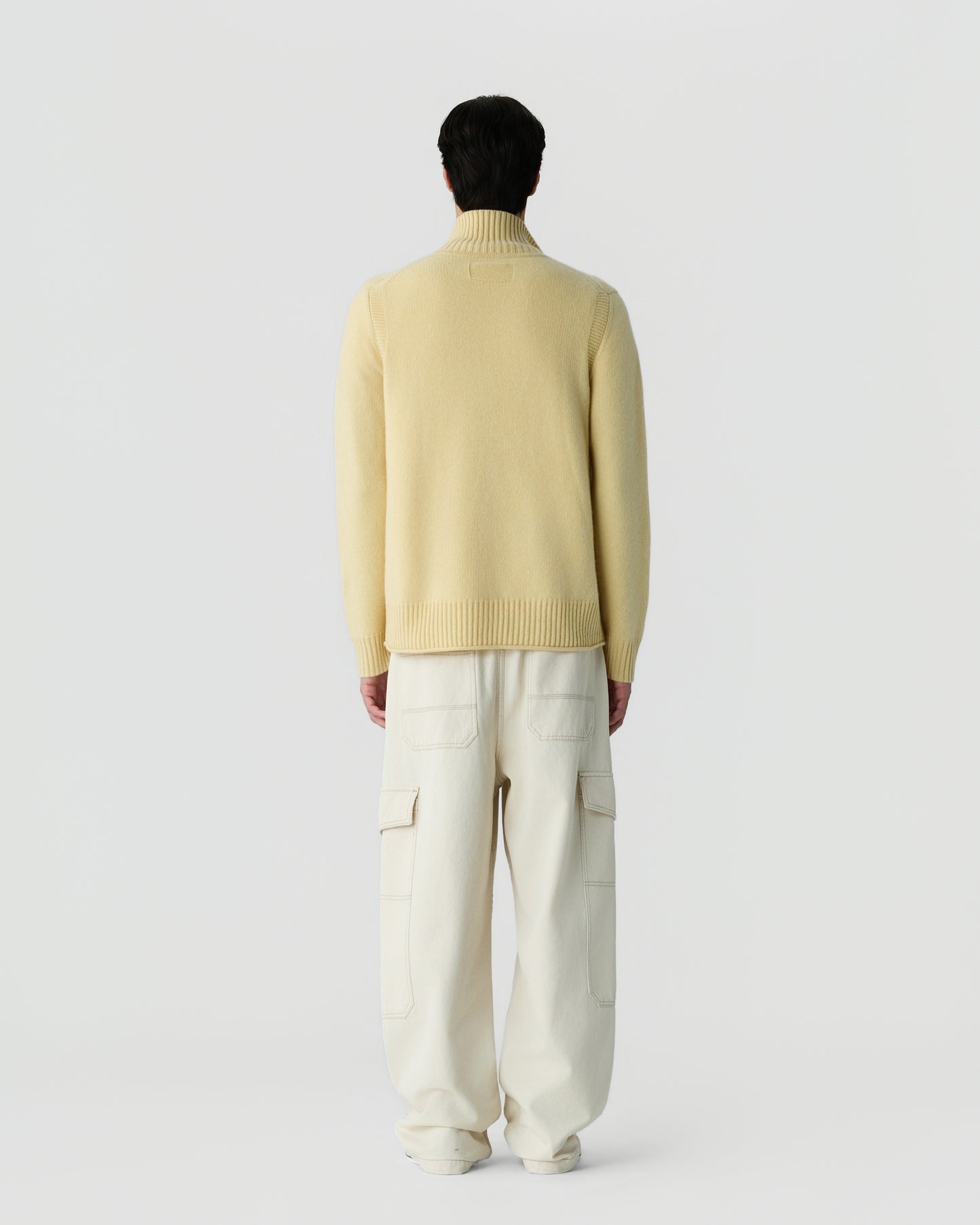 Rear view of a person wearing a light yellow cashmere turtleneck sweater paired with loose-fitting off-white cargo pants against a plain white background.