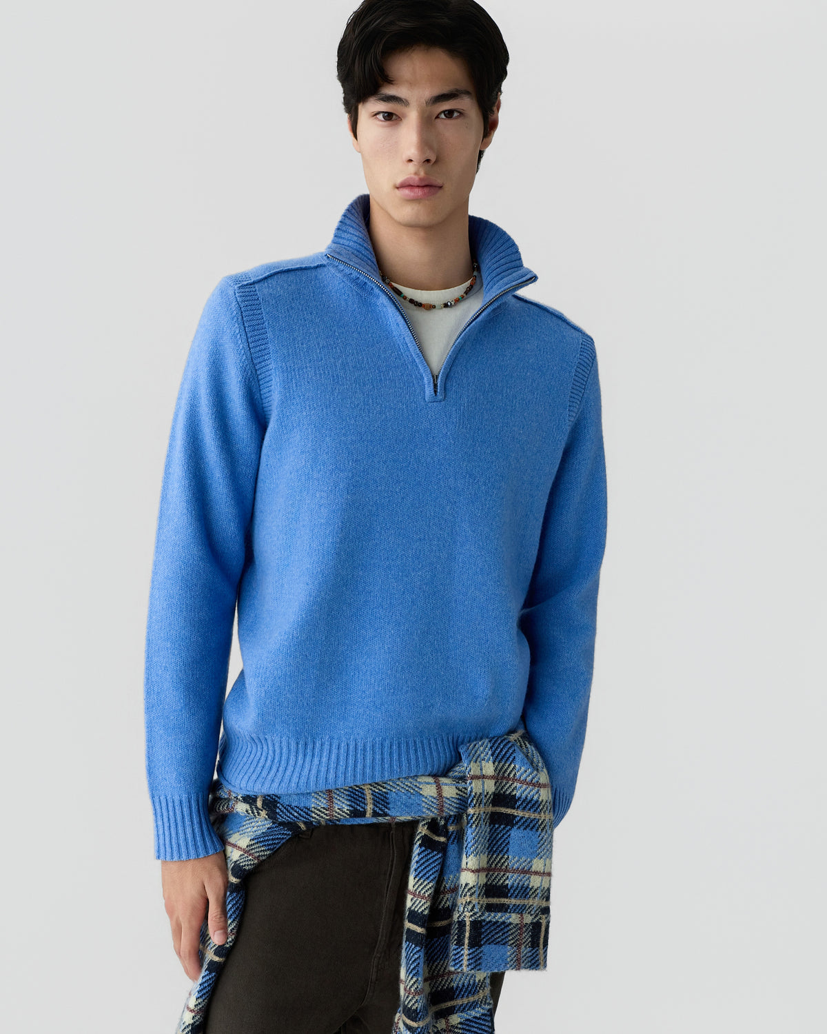 Young man wearing a light blue quarter-zip sweater with a ribbed collar, cuffs, and hem, standing against a plain light gray background with a plaid shirt tied around his waist.