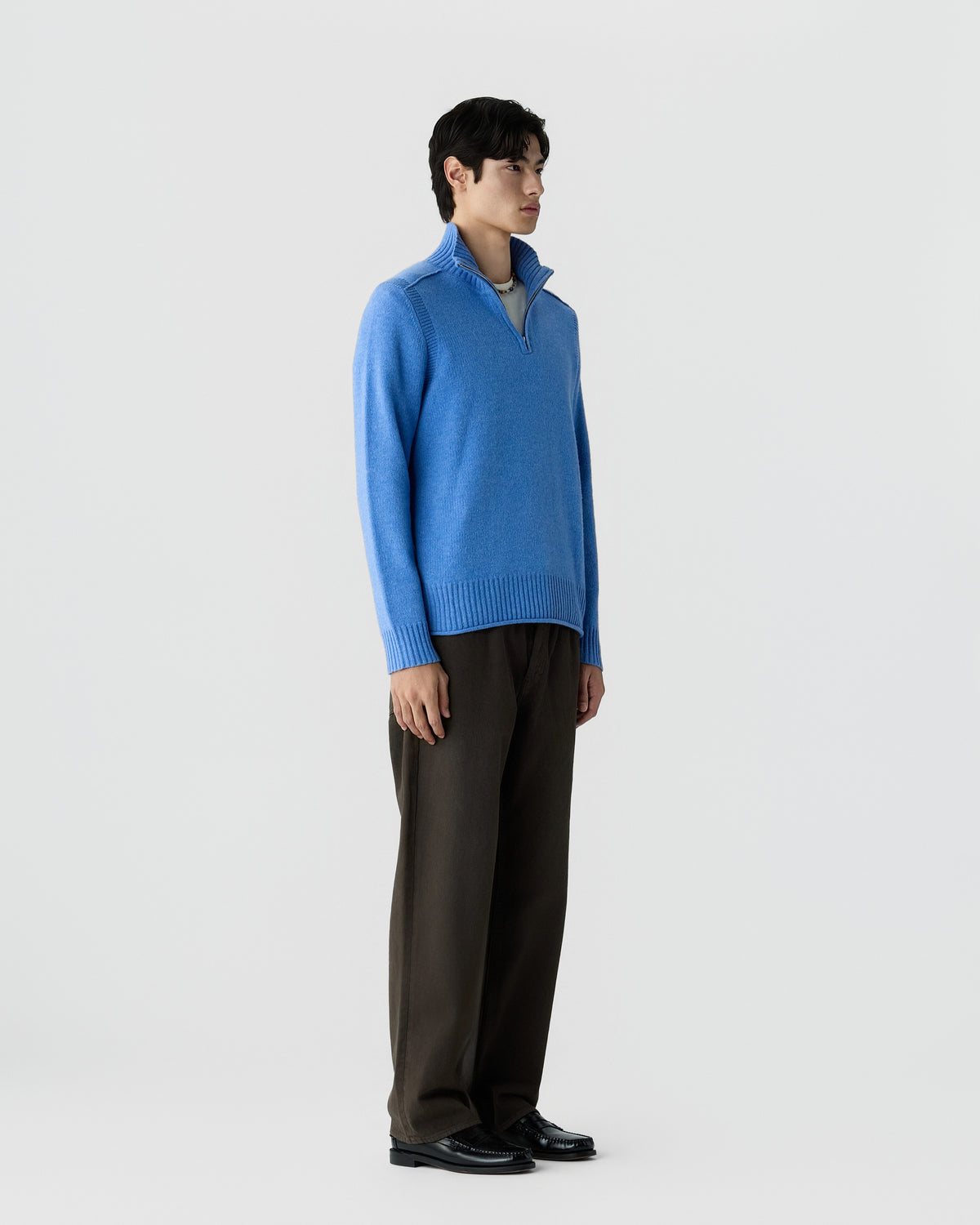 Young man standing wearing a blue half-zip sweater with ribbed cuffs and hem, dark wide-leg pants, and black dress shoes against a plain white background.