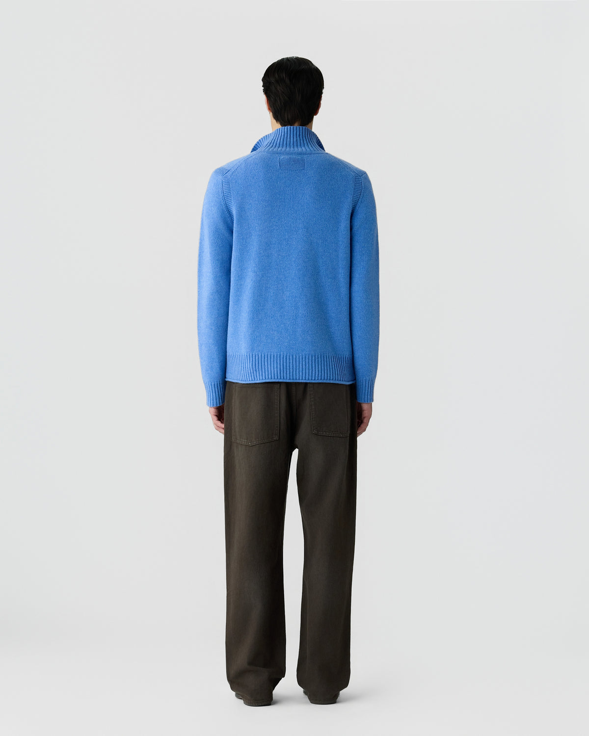 Man shown from the back wearing a bright blue turtleneck sweater and loose dark brown pants against a plain light gray background.