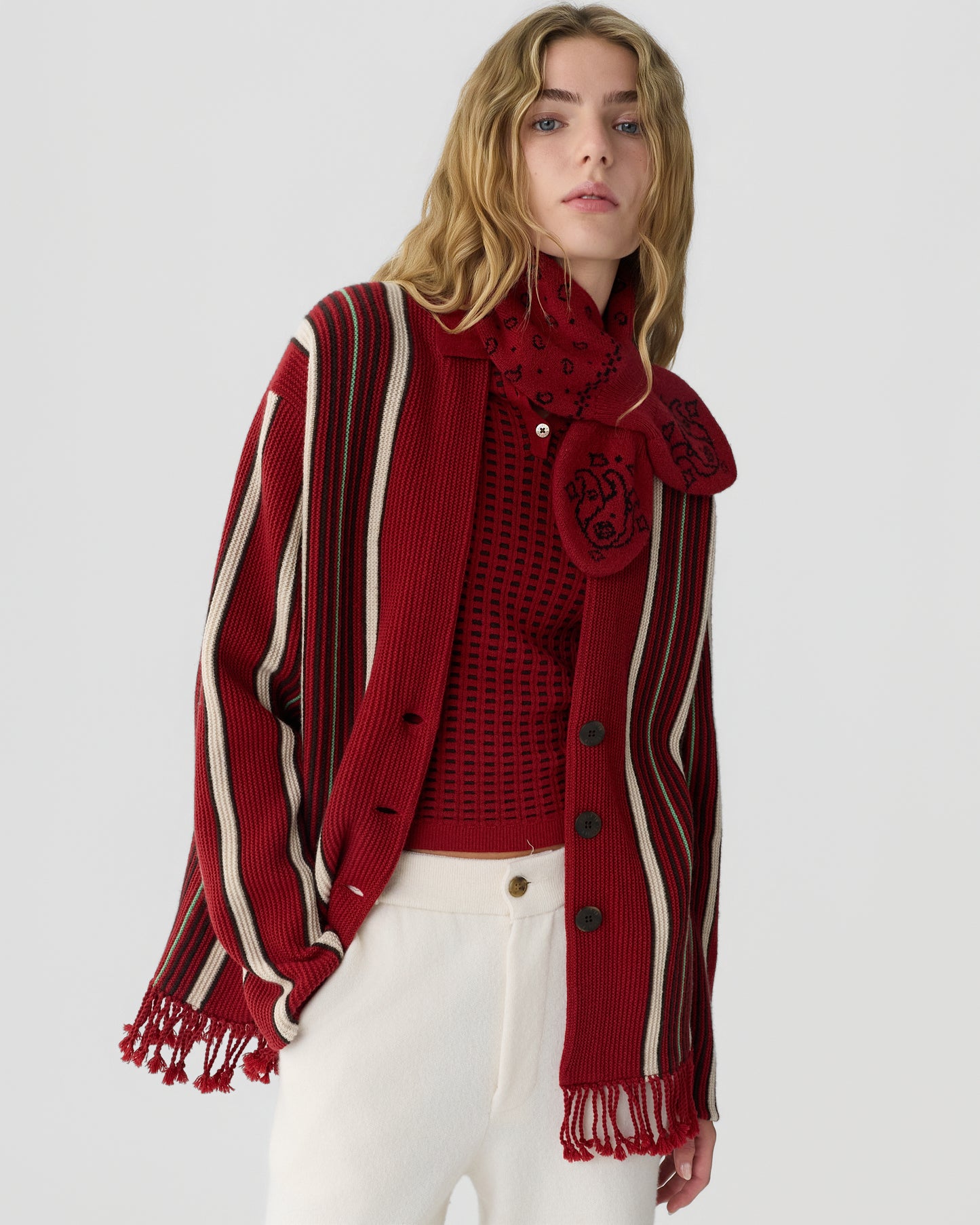 Woman wearing a red and cream striped cardigan with fringe details over a red patterned top and white pants, posing against a plain light background.