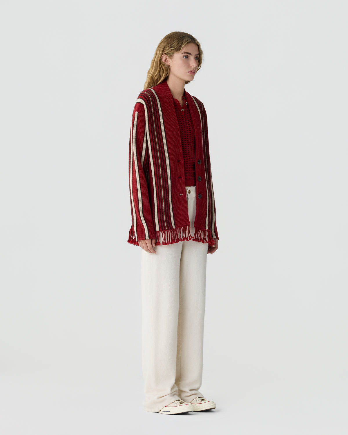 Young woman with blonde hair wearing a red and white striped knit cardigan over a red sweater, paired with loose white pants and white sneakers, standing against a plain background.