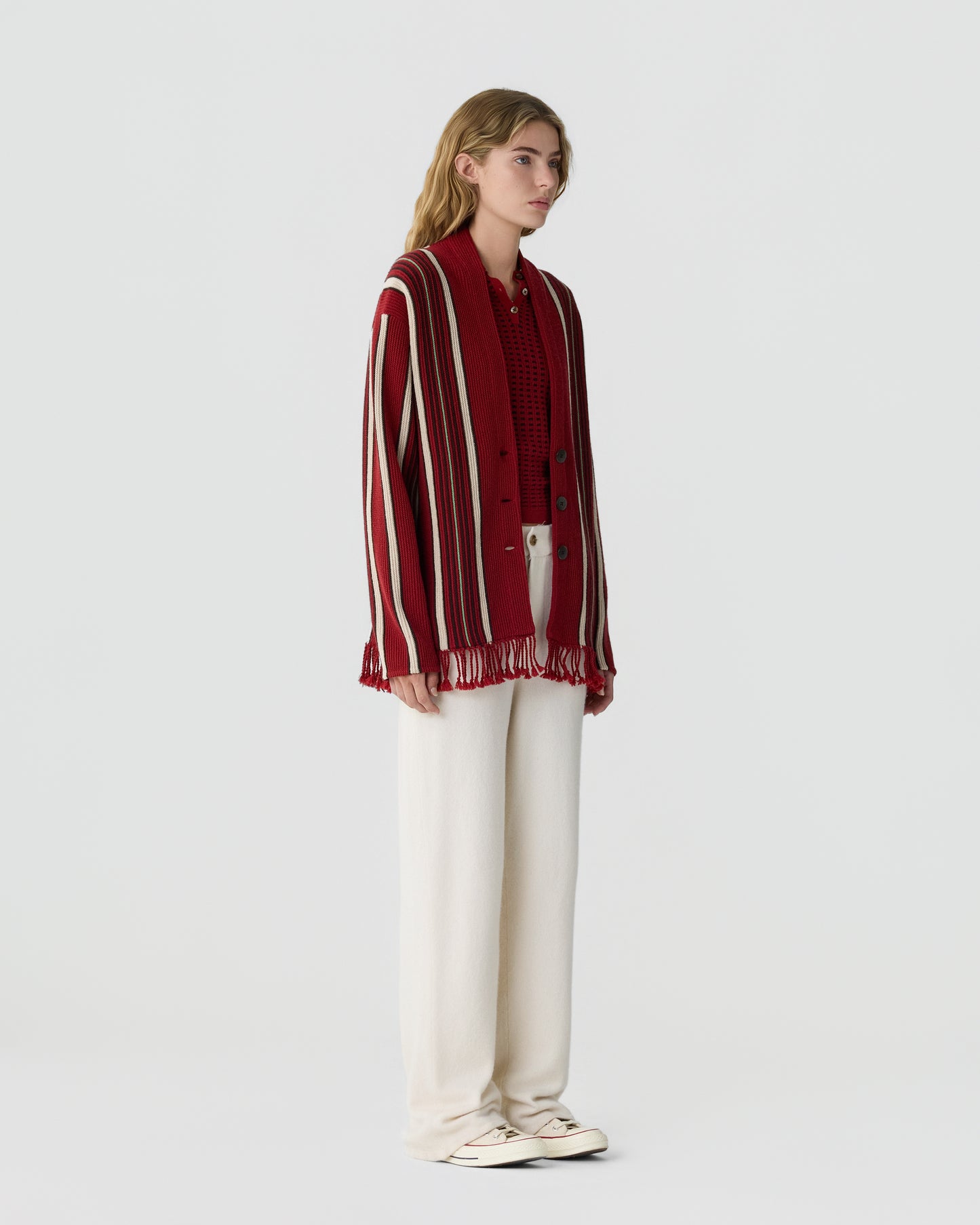 Young woman with blonde hair wearing a red and white striped knit cardigan over a red sweater, paired with loose white pants and white sneakers, standing against a plain background.