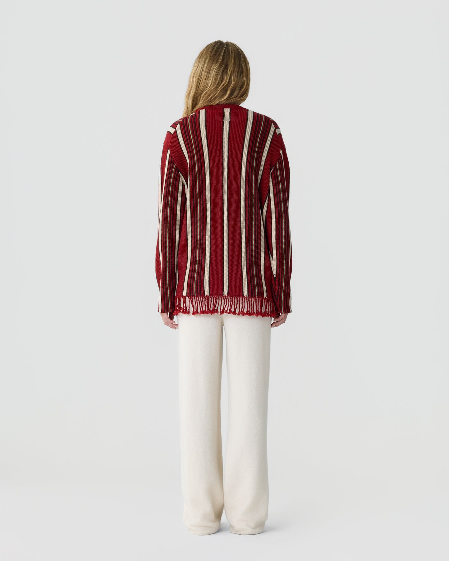 Woman wearing a long-sleeve red sweater with vertical beige and black stripes and fringe at the hem, paired with loose white pants, standing facing away against a plain background.