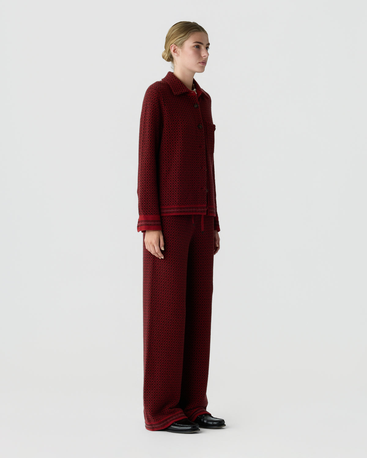 Woman standing in profile wearing a burgundy patterned shirt with matching wide leg trousers and black shoes against a light gray background