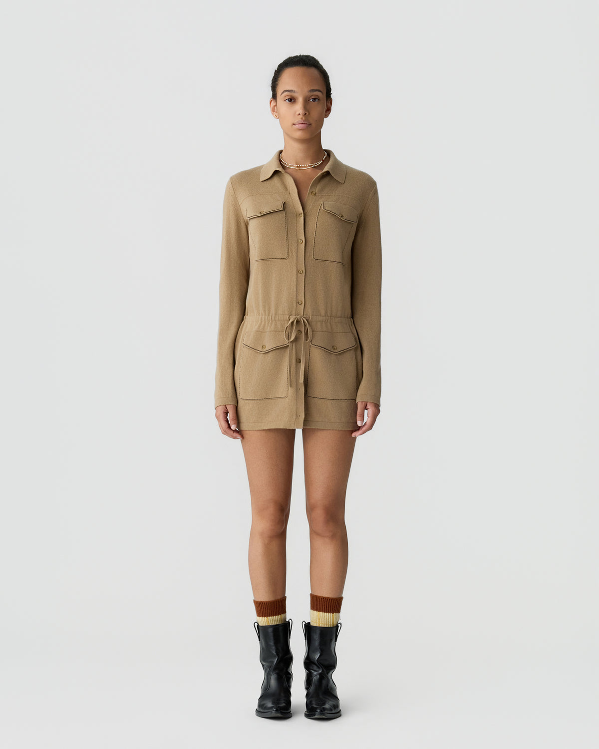 Woman standing against a plain light gray background wearing a long-sleeve tan shirt dress with front pockets, a drawstring waist, brown and mustard socks, and black ankle boots.