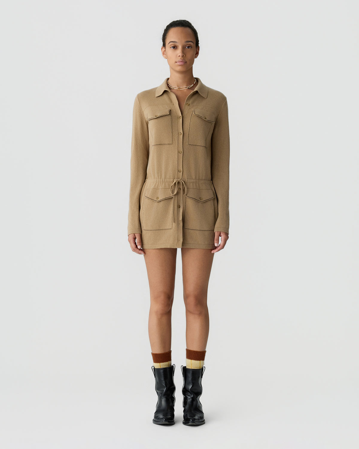 Model standing against a light gray background wearing a tan long sleeve utility romper with chest pockets and black knee high boots.