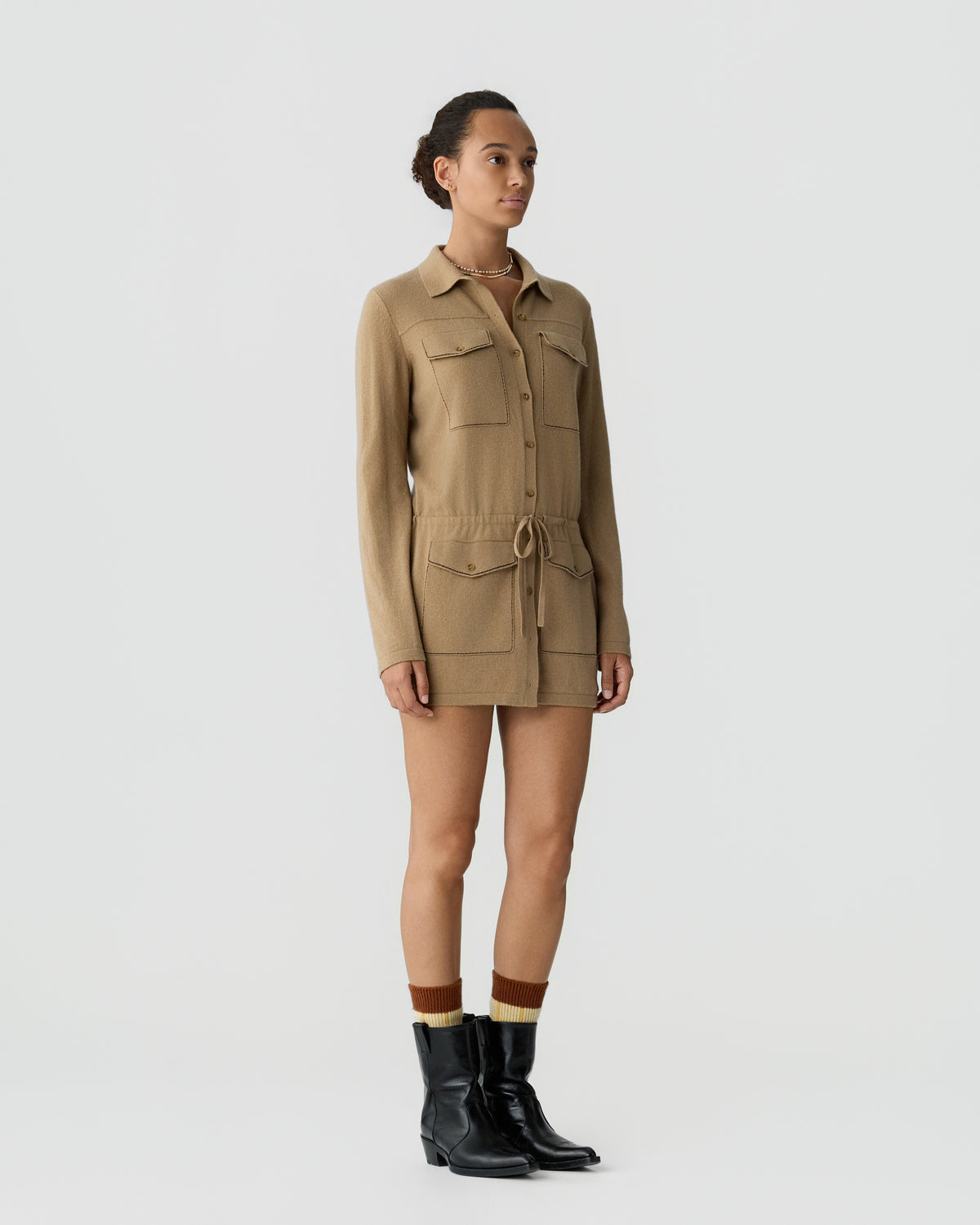 Woman wearing a long-sleeve beige button-up dress with front pockets and a drawstring waist, paired with black ankle boots and striped socks against a plain background.