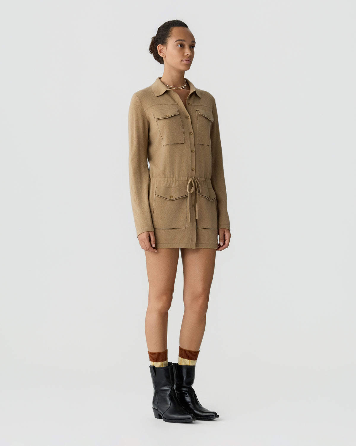 Model wearing a tan long sleeve utility jumpsuit with chest pockets and a drawstring waist, paired with brown socks and black ankle boots against a light background