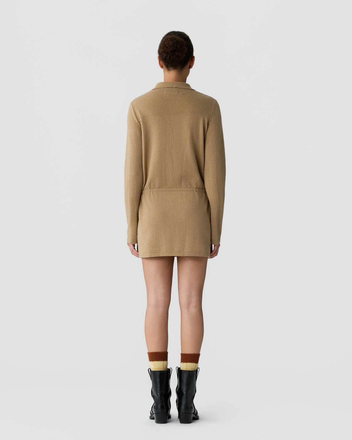 Rear view of a person wearing a long-sleeve, camel-colored knit dress paired with contrasting two-tone socks and black ankle boots against a plain white background.