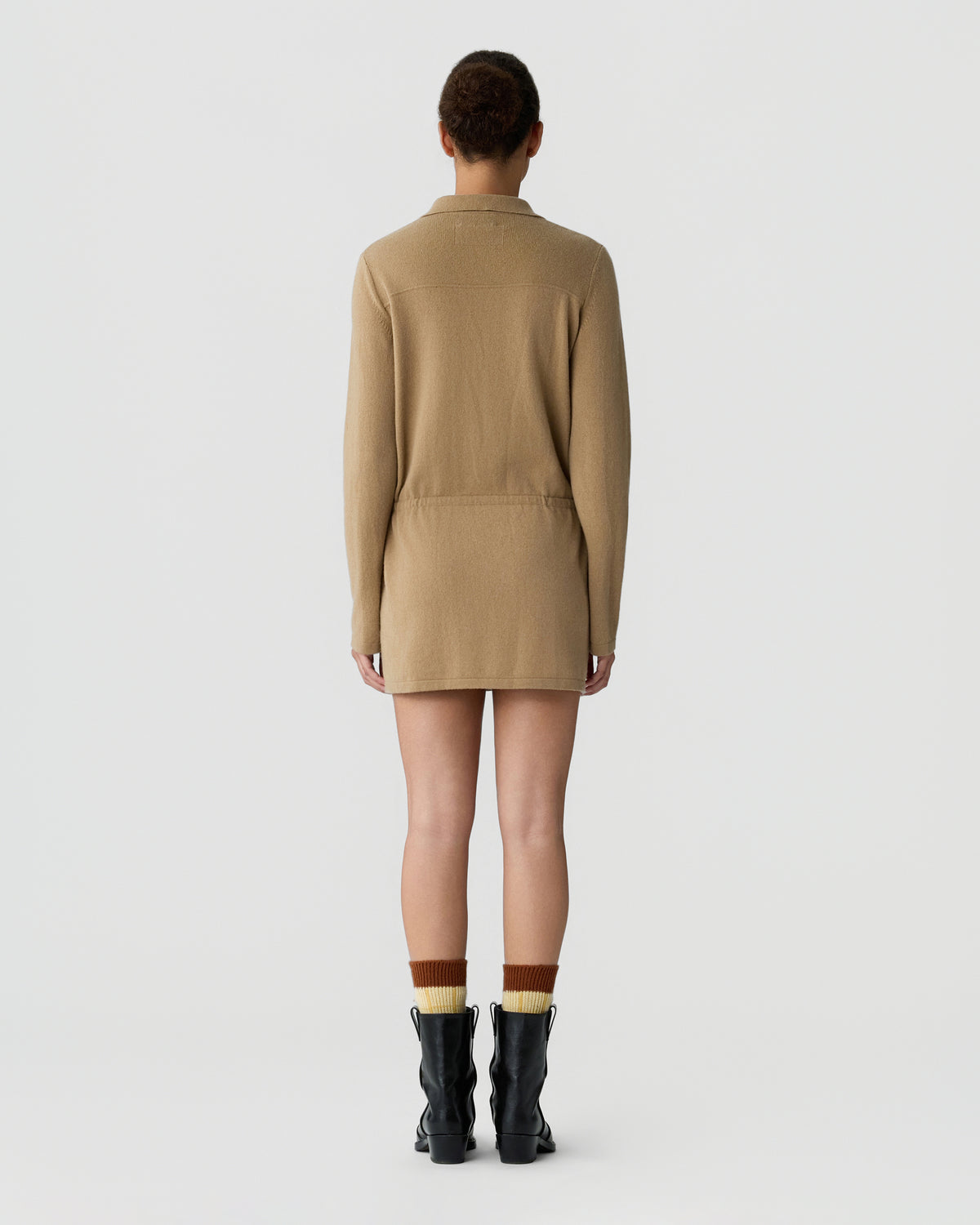Back view of a person wearing a tan long sleeve knit dress with a waist seam, brown socks, and black ankle boots against a light gray background