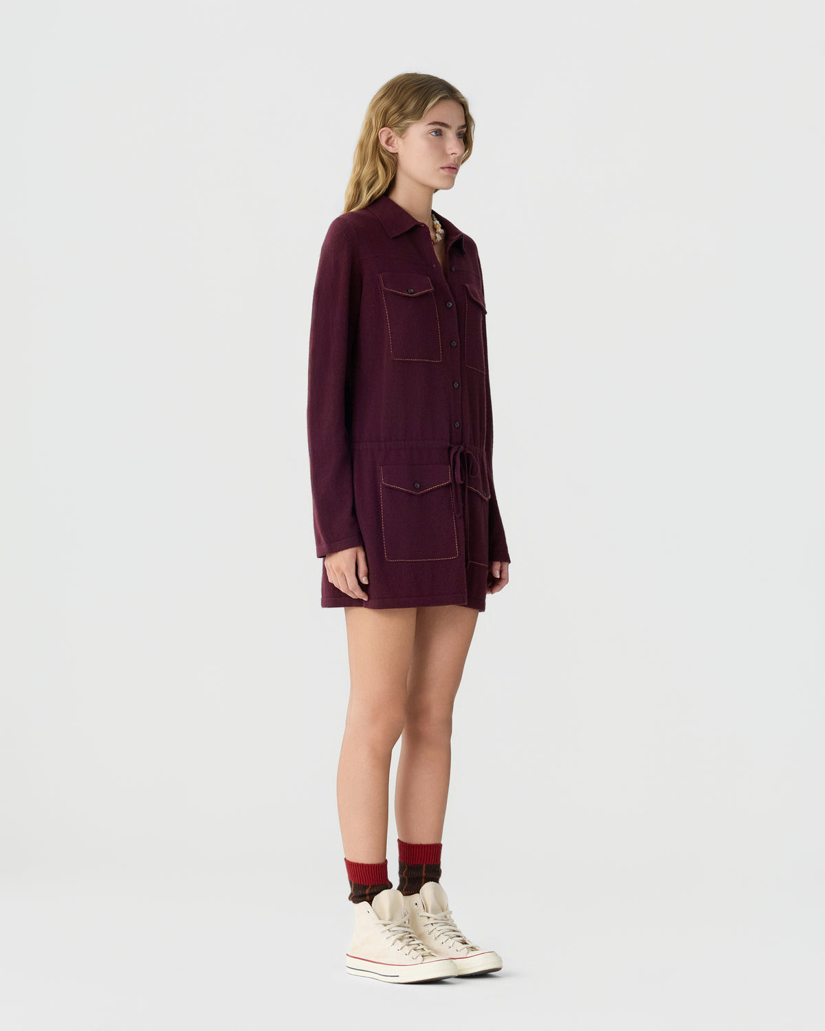 Young woman standing in profile wearing a long-sleeve, button-up burgundy shirt dress with front pockets, paired with two-tone socks and high-top sneakers against a plain background.