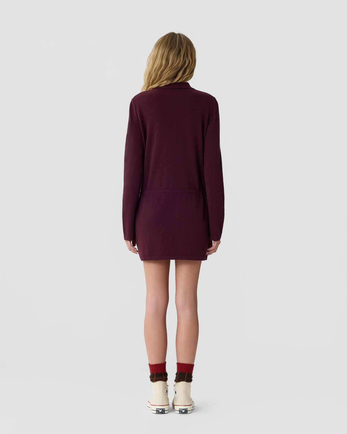 Woman with long blonde hair wearing a maroon long-sleeve dress standing facing away on a plain light background with white boots and red socks.