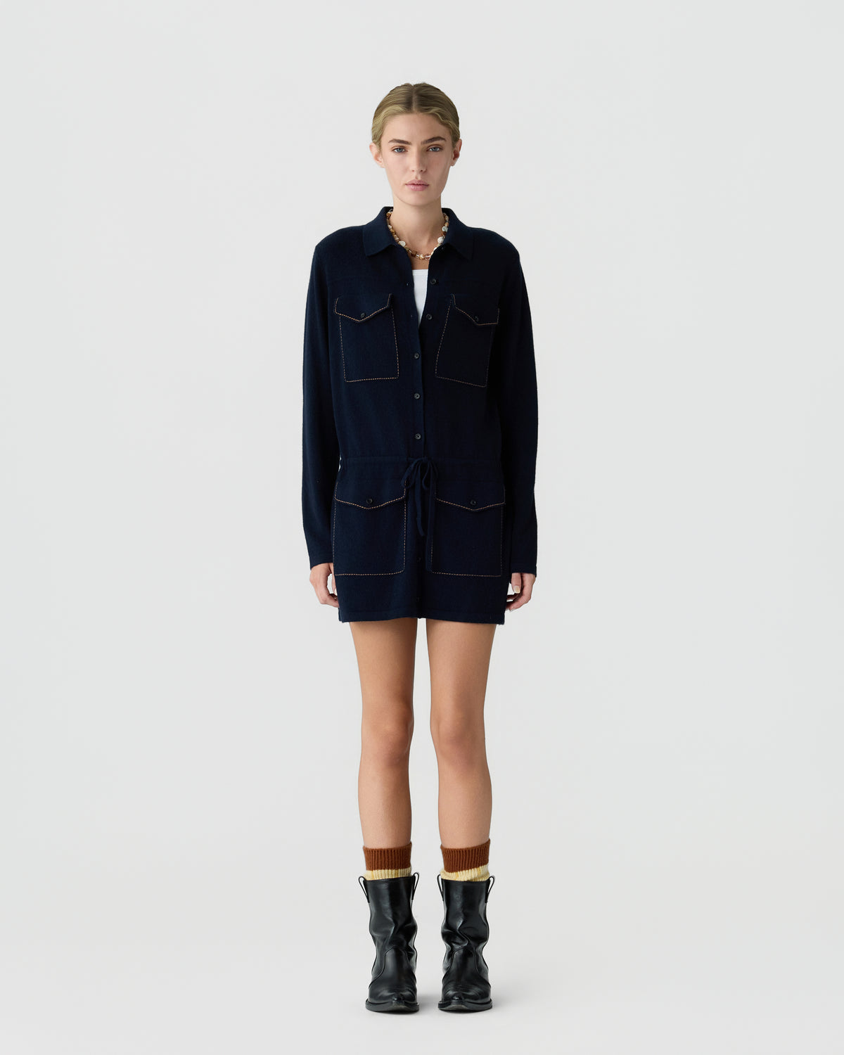Young woman standing against a plain white background wearing a navy long-sleeve buttoned dress with patch pockets, brown socks, and black ankle boots.