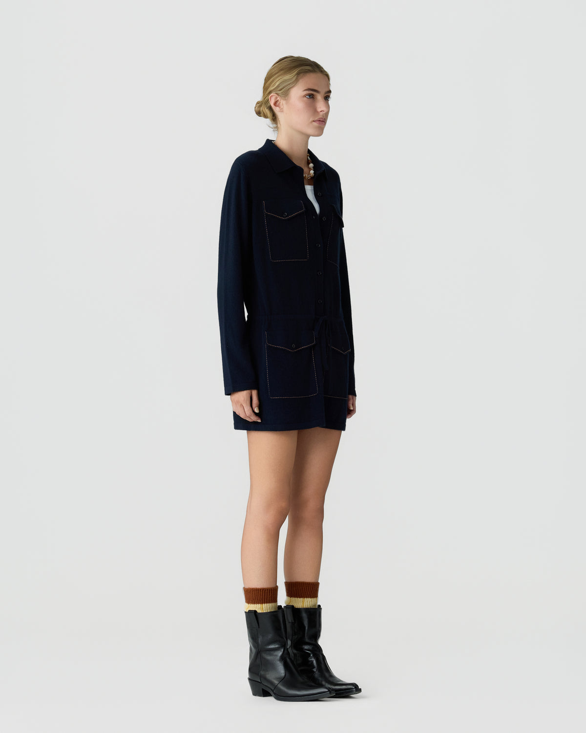 Woman standing sideways wearing a navy long-sleeve jacket dress with four front pockets, a white top underneath, multicolored socks, and black ankle boots against a plain white background.