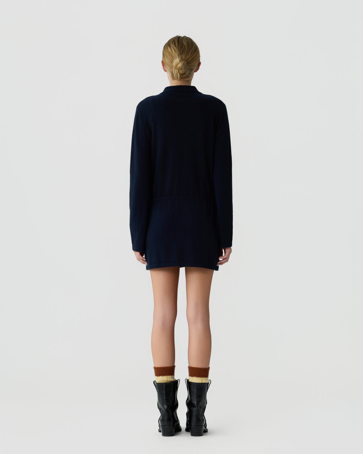 Rear view of a person standing against a plain background wearing a long-sleeve dark navy garment, brown and beige socks, and black boots, with hair tied back.
