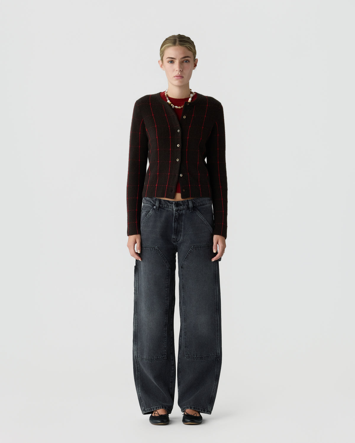 A woman stands facing forward wearing a dark fitted cardigan with thin red vertical stripes and wide-leg faded black jeans, paired with black shoes and a white beaded necklace.