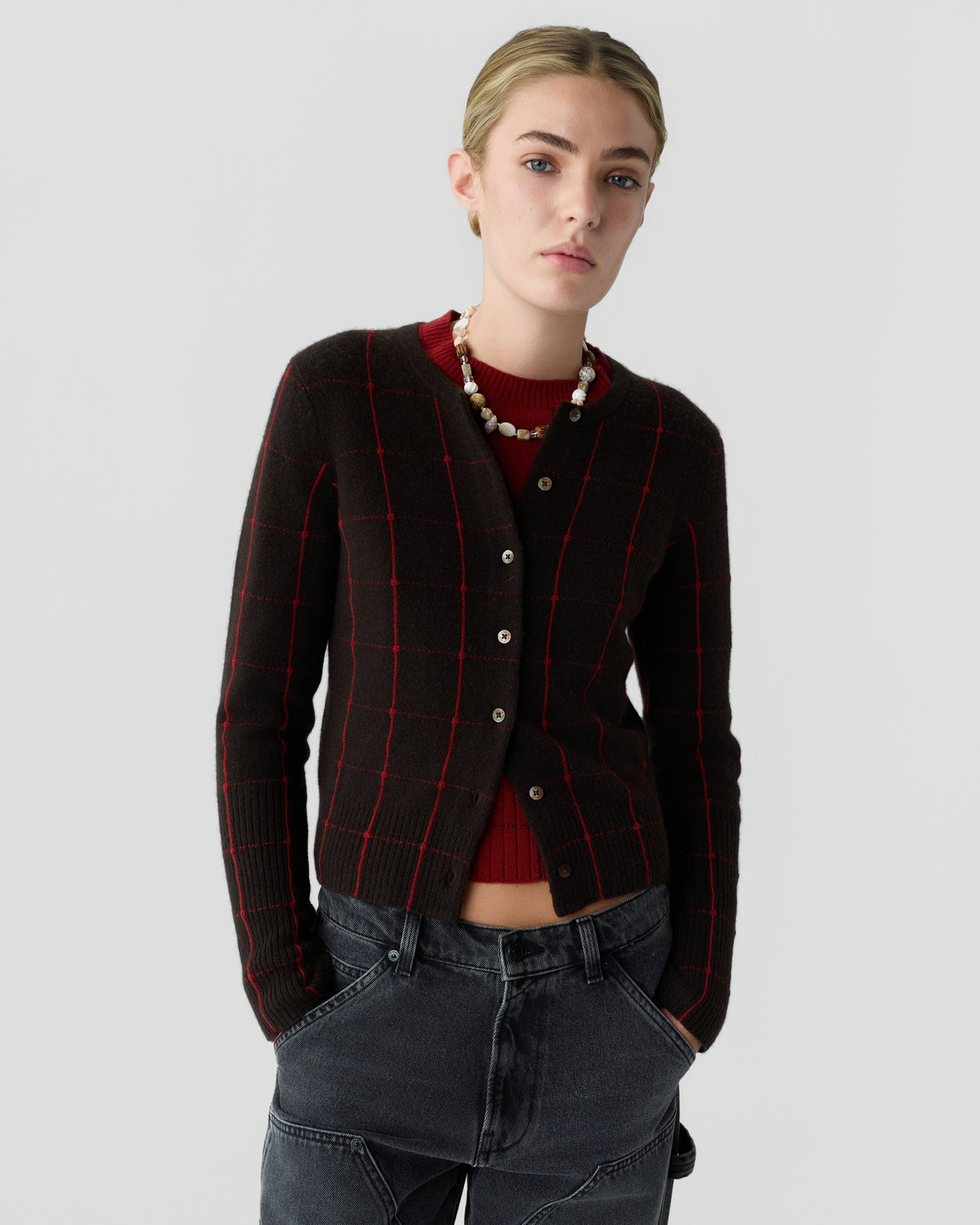 Young woman wearing a black cardigan with red windowpane pattern over a red sweater, paired with high-waisted dark gray jeans, standing against a plain light gray background.