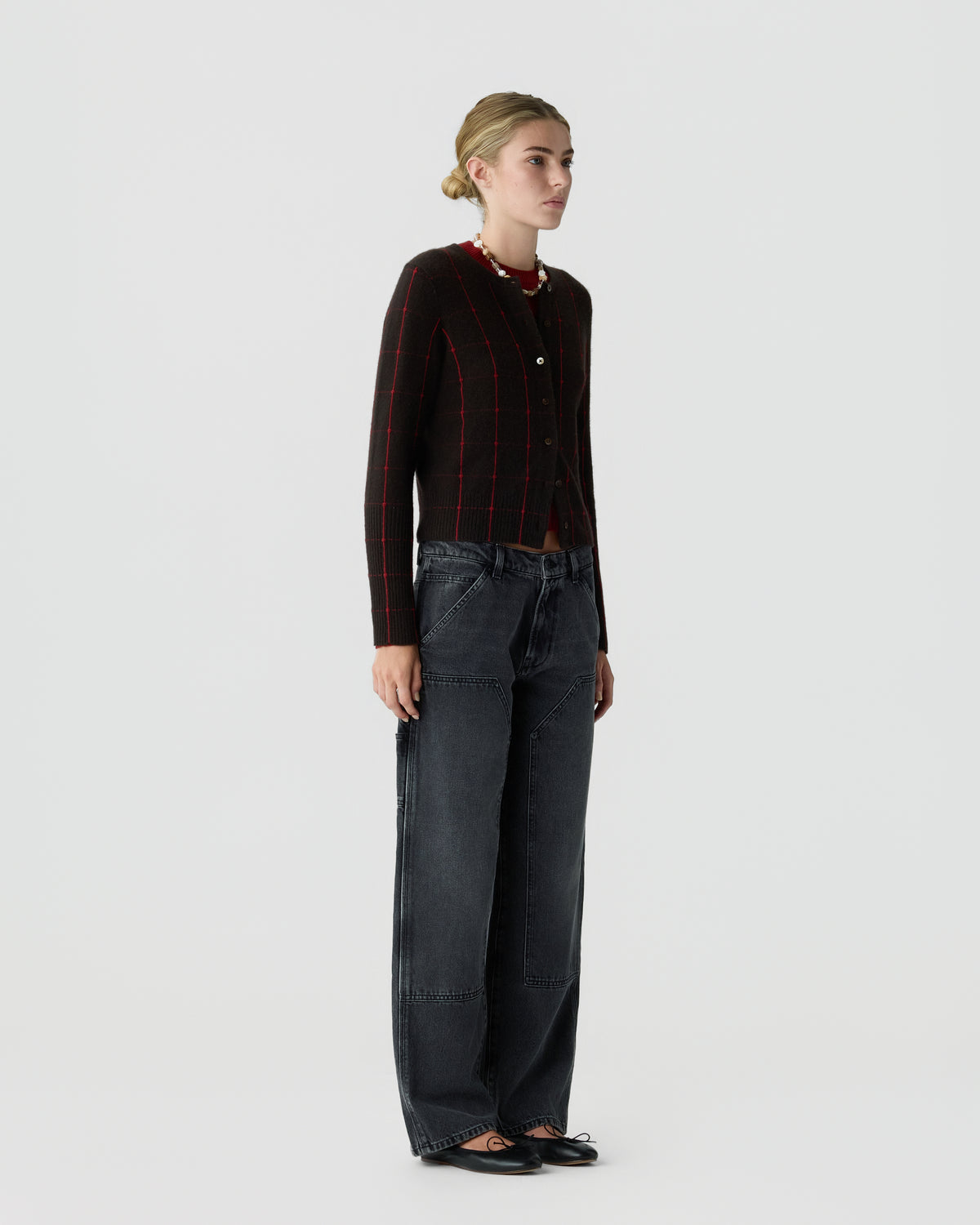 Woman standing against a plain background wearing a dark brown cardigan with red lines, black wide-leg pants with patch details, a beaded necklace, and black ballet flats.