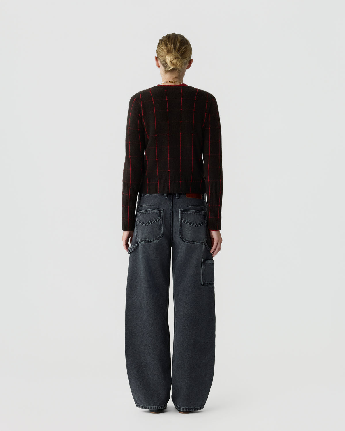 Rear view of a person wearing a black long-sleeve top with red grid lines and loose-fitting dark gray jeans with patch pockets against a plain light background.