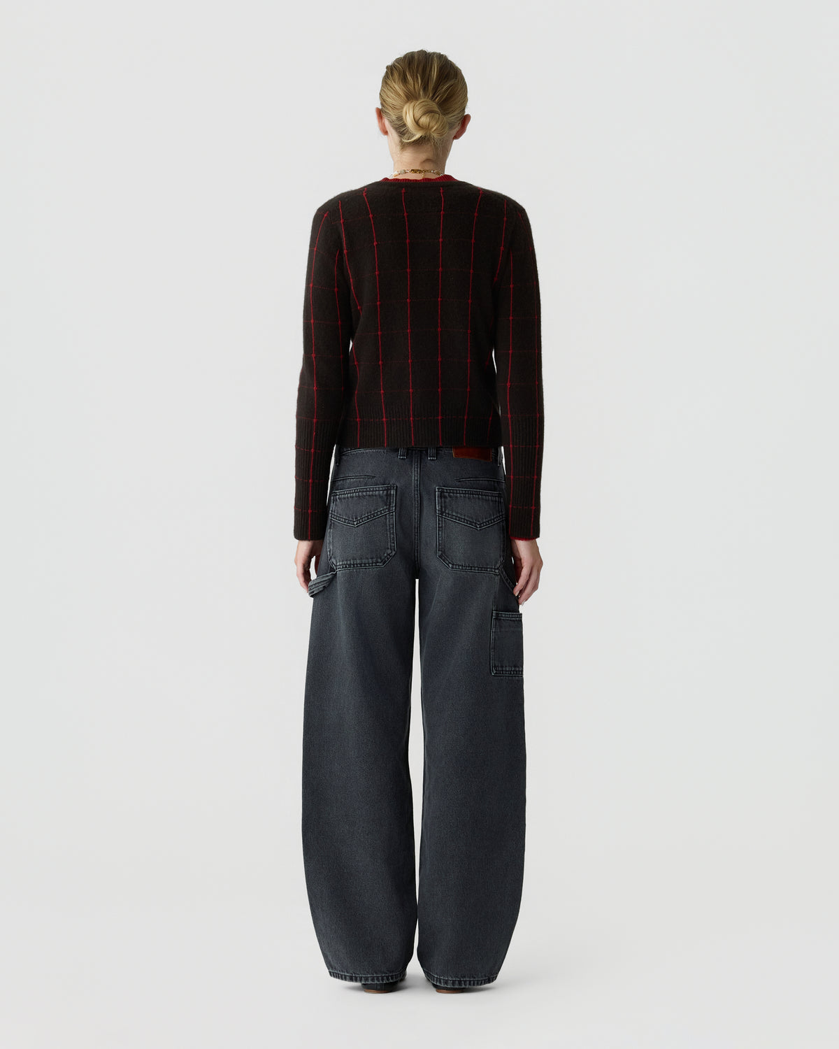 Back view of a person wearing a black and red checked sweater with wide leg dark gray cargo jeans and blonde hair tied in a bun against a light background