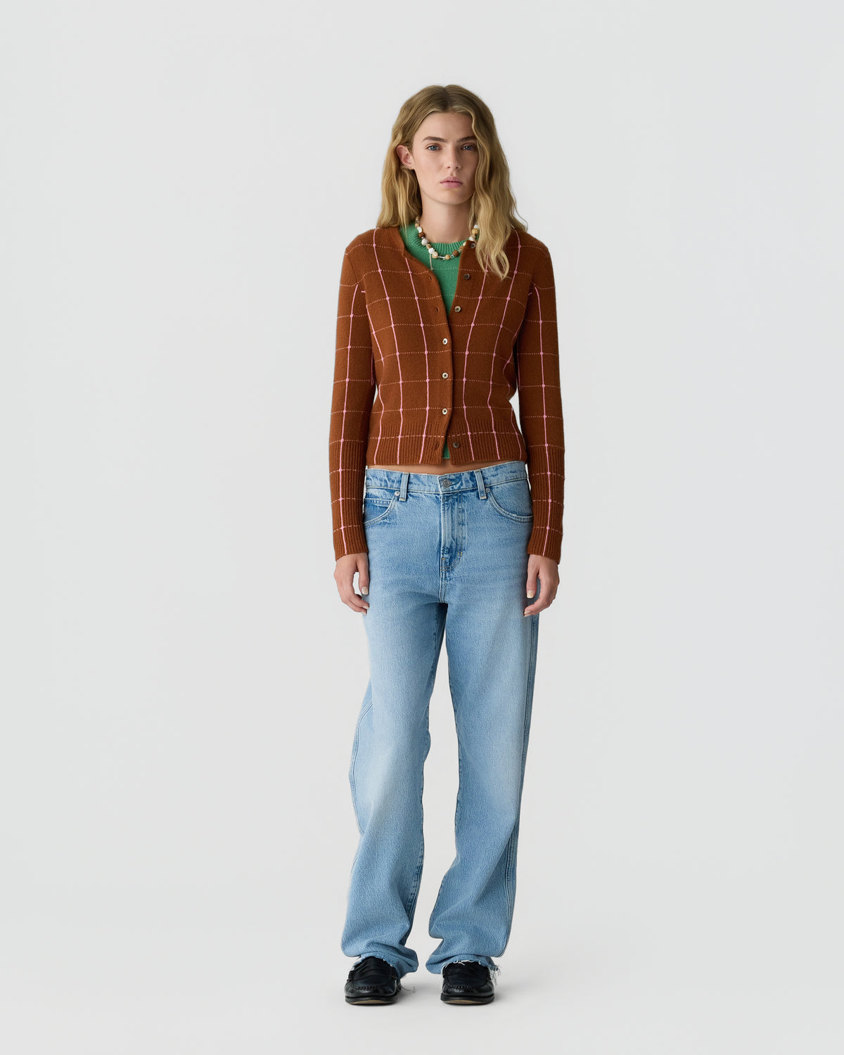Young woman with long blonde hair wearing a rust-colored buttoned cardigan with a white check pattern, green shirt underneath, light blue jeans, and black shoes standing against a plain white background.