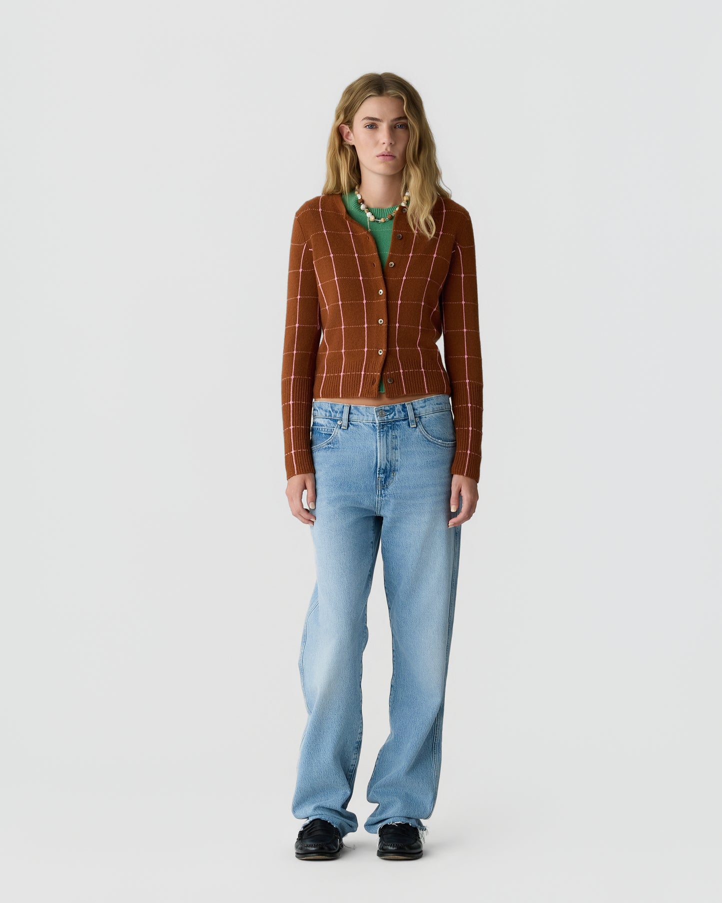 Young woman with long blonde hair wearing a rust-colored buttoned cardigan with a white check pattern, green shirt underneath, light blue jeans, and black shoes standing against a plain white background.