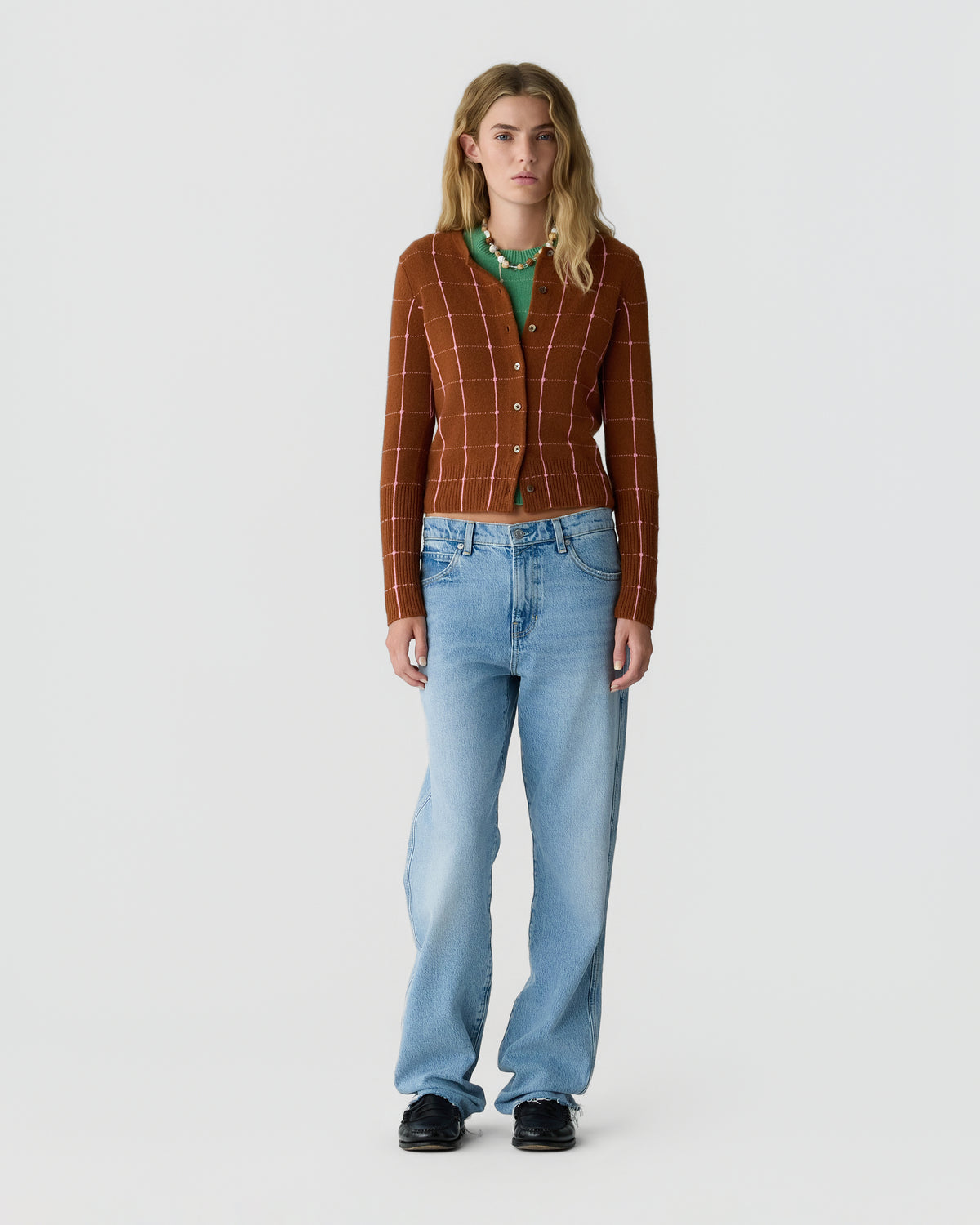 Full-length portrait of a woman wearing a brown checked cardigan over a green top with light blue jeans and black shoes against a pale gray background