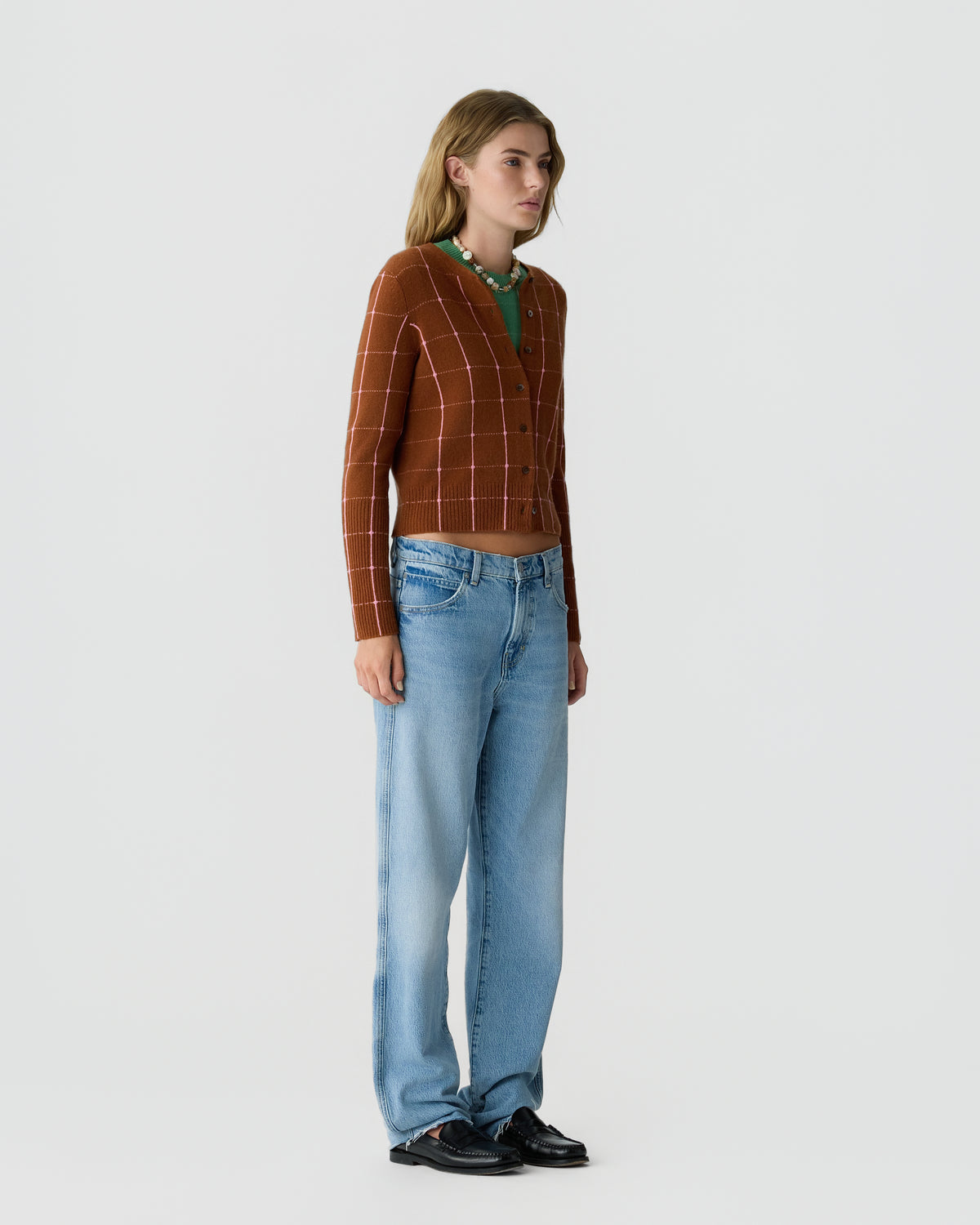 A woman standing against a plain background wearing a brown checkered cardigan over a green top, light blue high-waisted jeans, and black shoes.