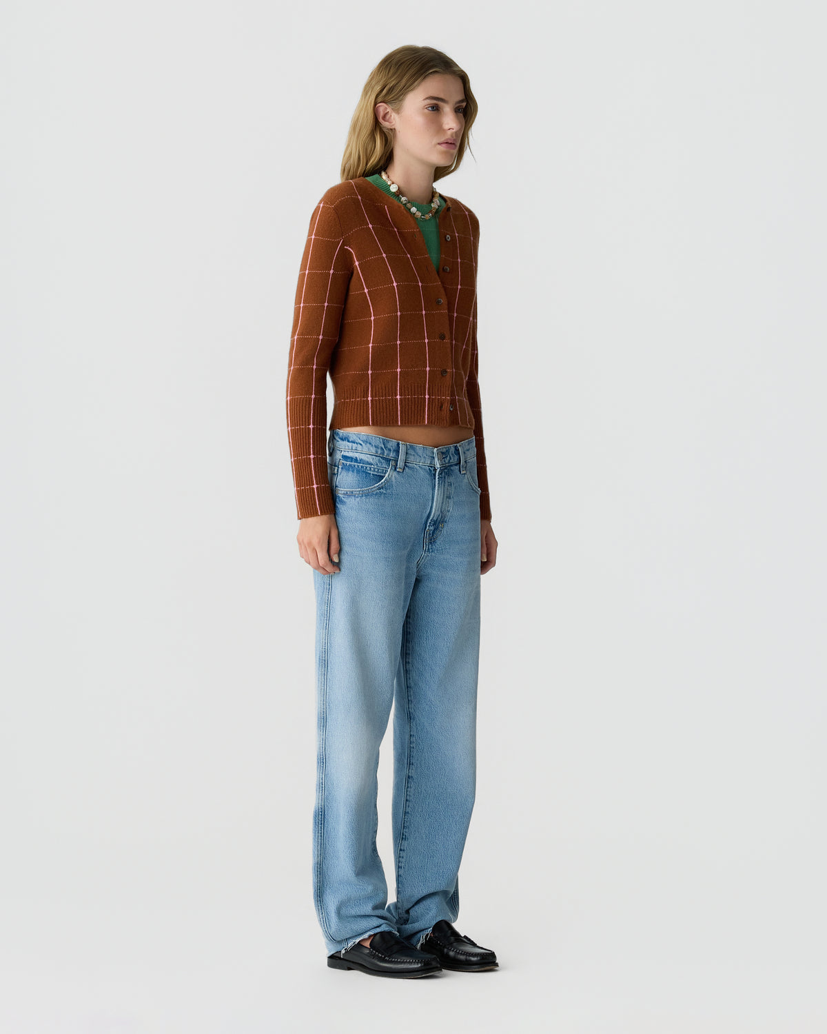 Model wearing a brown checked cardigan with a green collar over light blue wide leg jeans and black shoes against a pale gray background