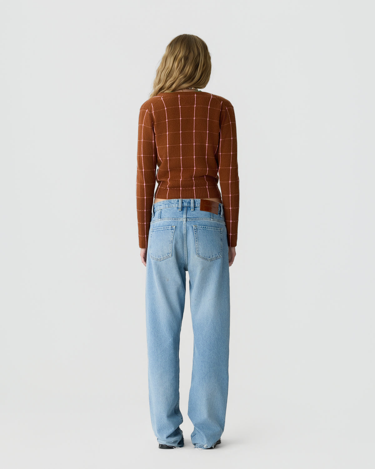 Woman standing facing away wearing a rust-colored sweater with a white grid pattern and light blue high-waisted jeans against a plain light background.