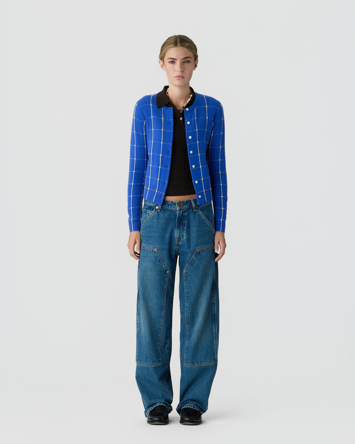 Front-facing model wearing a blue checked button cardigan over a black top, with relaxed blue jeans and black shoes against a light gray background