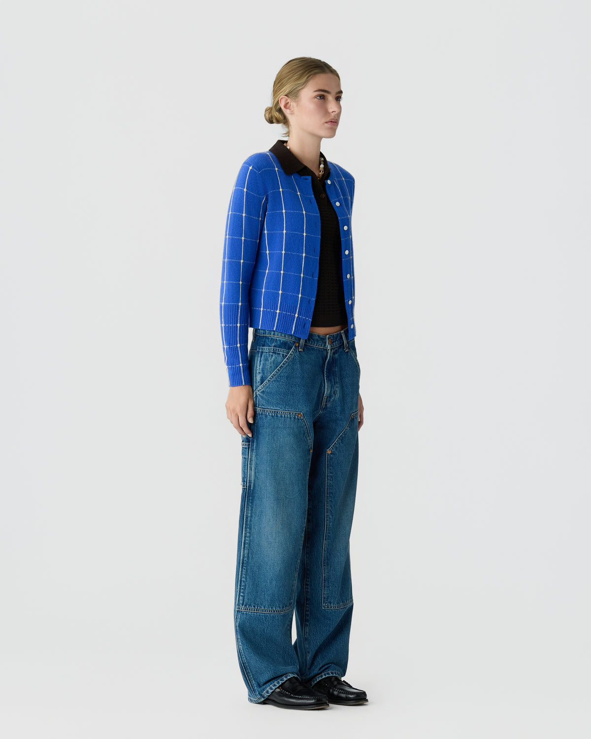 Woman wearing a blue checked button front cardigan with a dark shirt loose blue jeans and black shoes against a light gray background