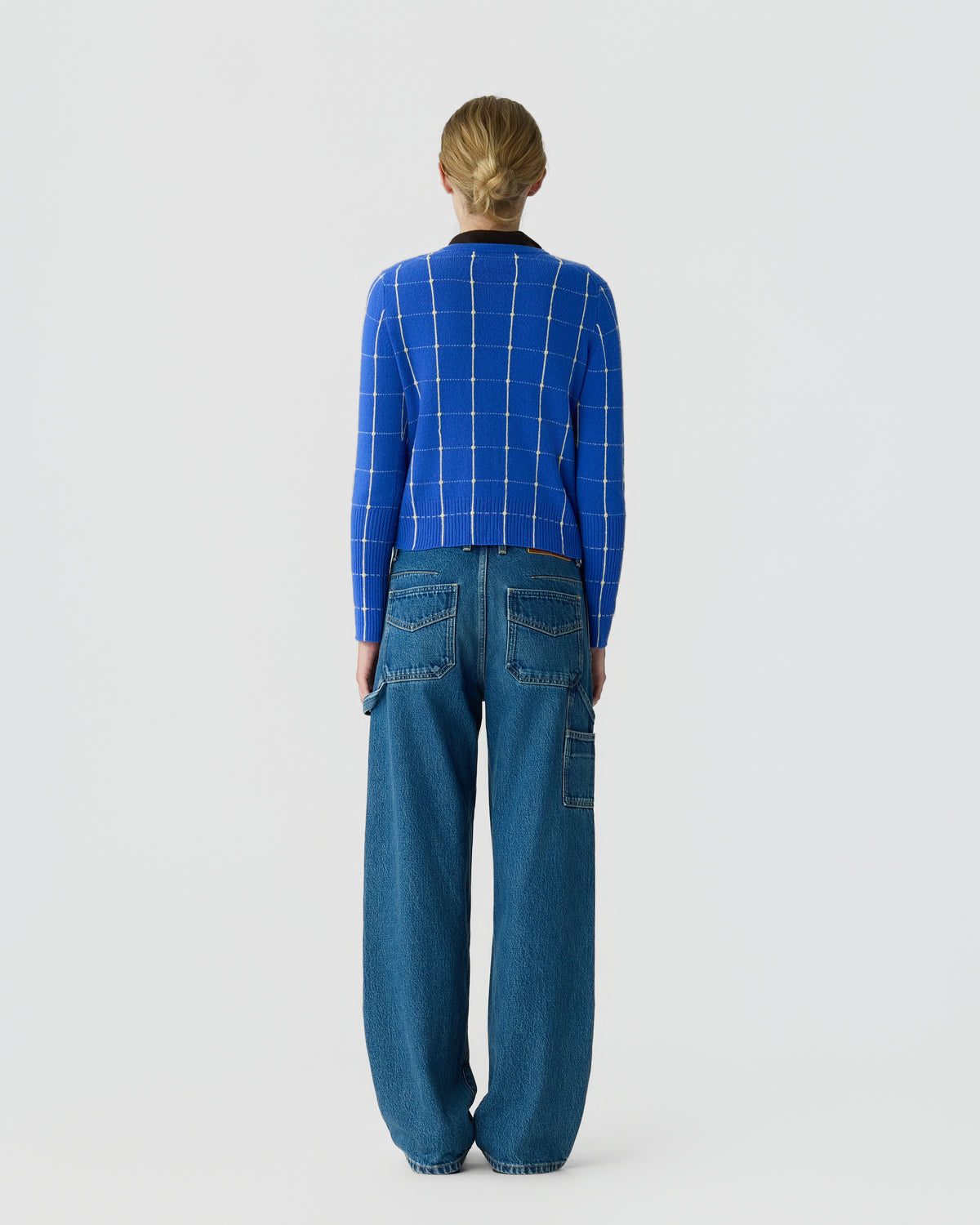 Back view of a person wearing a blue windowpane check sweater and relaxed blue jeans against a plain light gray background