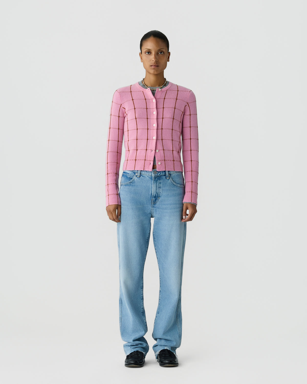 Person wearing a pink checked button front cardigan with blue jeans and black shoes against a light gray studio background